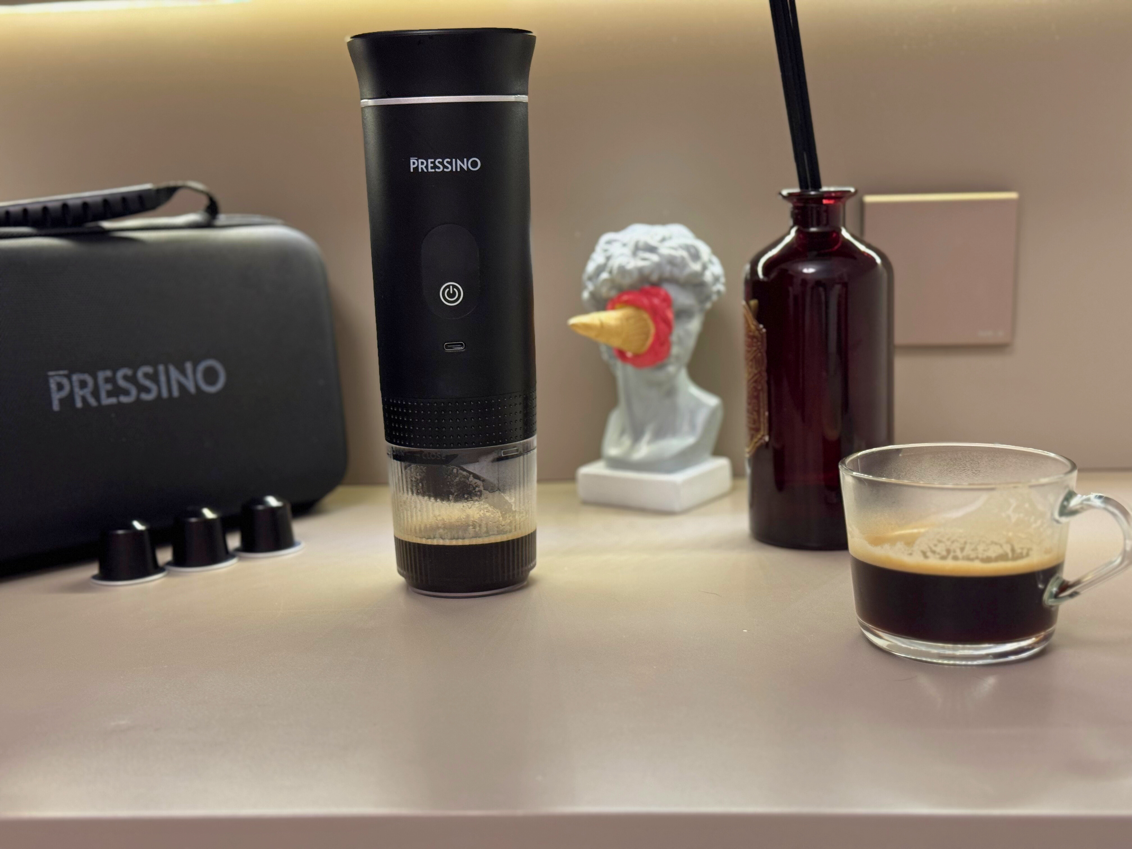 A black portable espresso maker labeled “Pressino” stands on a countertop beside a glass cup of freshly brewed espresso, a dark red bottle, a small decorative statue, and a Pressino-branded travel case with coffee capsules placed nearby. Warm lighting highlights the crema and modern setup.