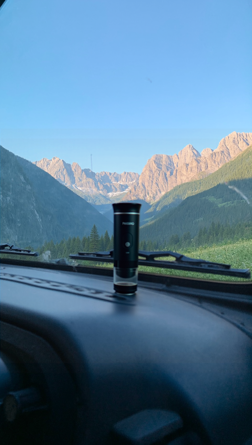 A black portable espresso maker labeled “PRESSINO” sits on a car dashboard overlooking a breathtaking mountain valley under a clear blue sky, blending the ritual of coffee with the freedom of the open road.