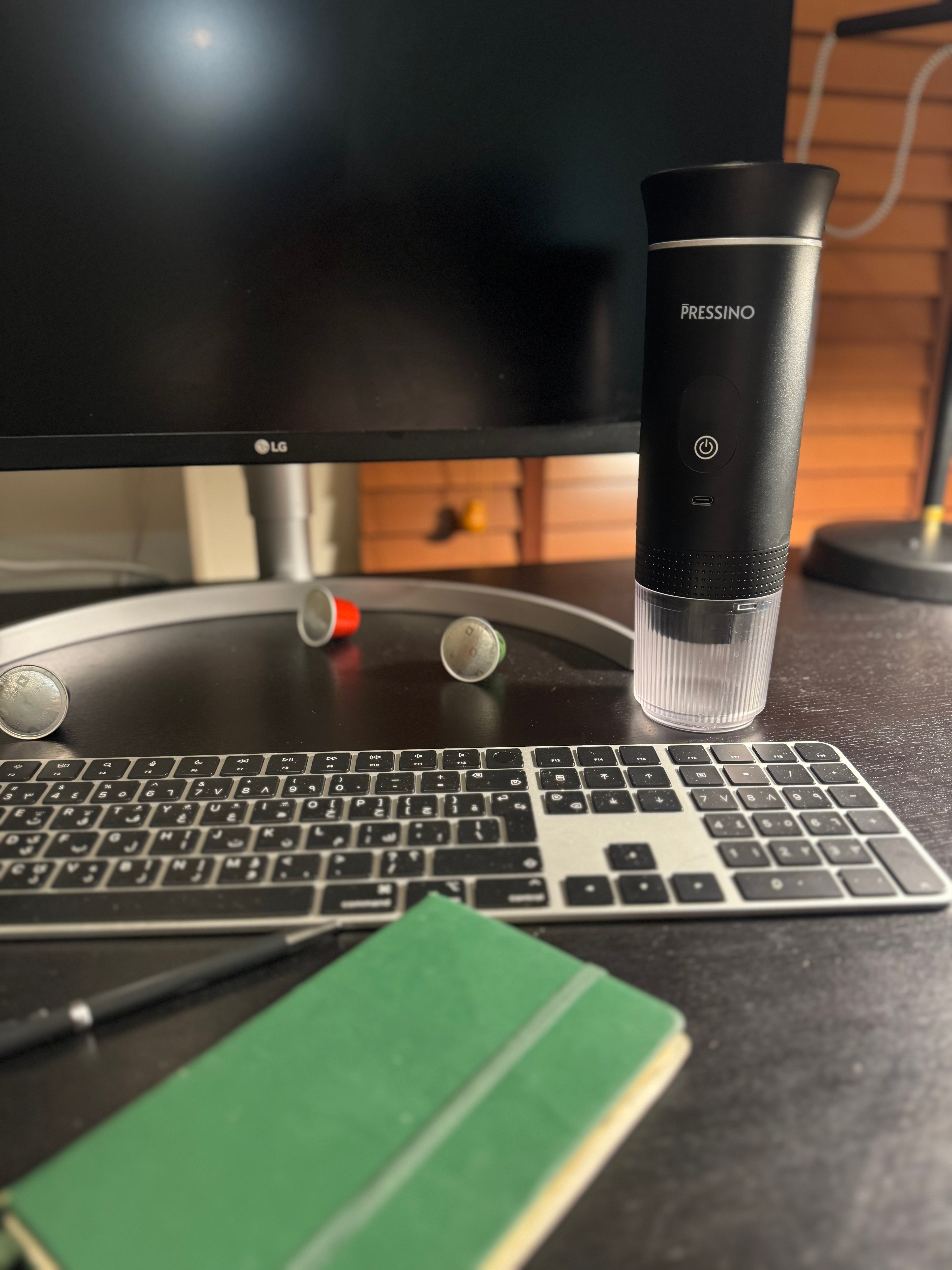 A black portable espresso maker labeled “PRESSINO” stands on a dark wooden desk beside a keyboard, green notebook, and scattered coffee capsules, with a blank monitor and warm lighting in the background—capturing a minimalist workspace coffee moment.