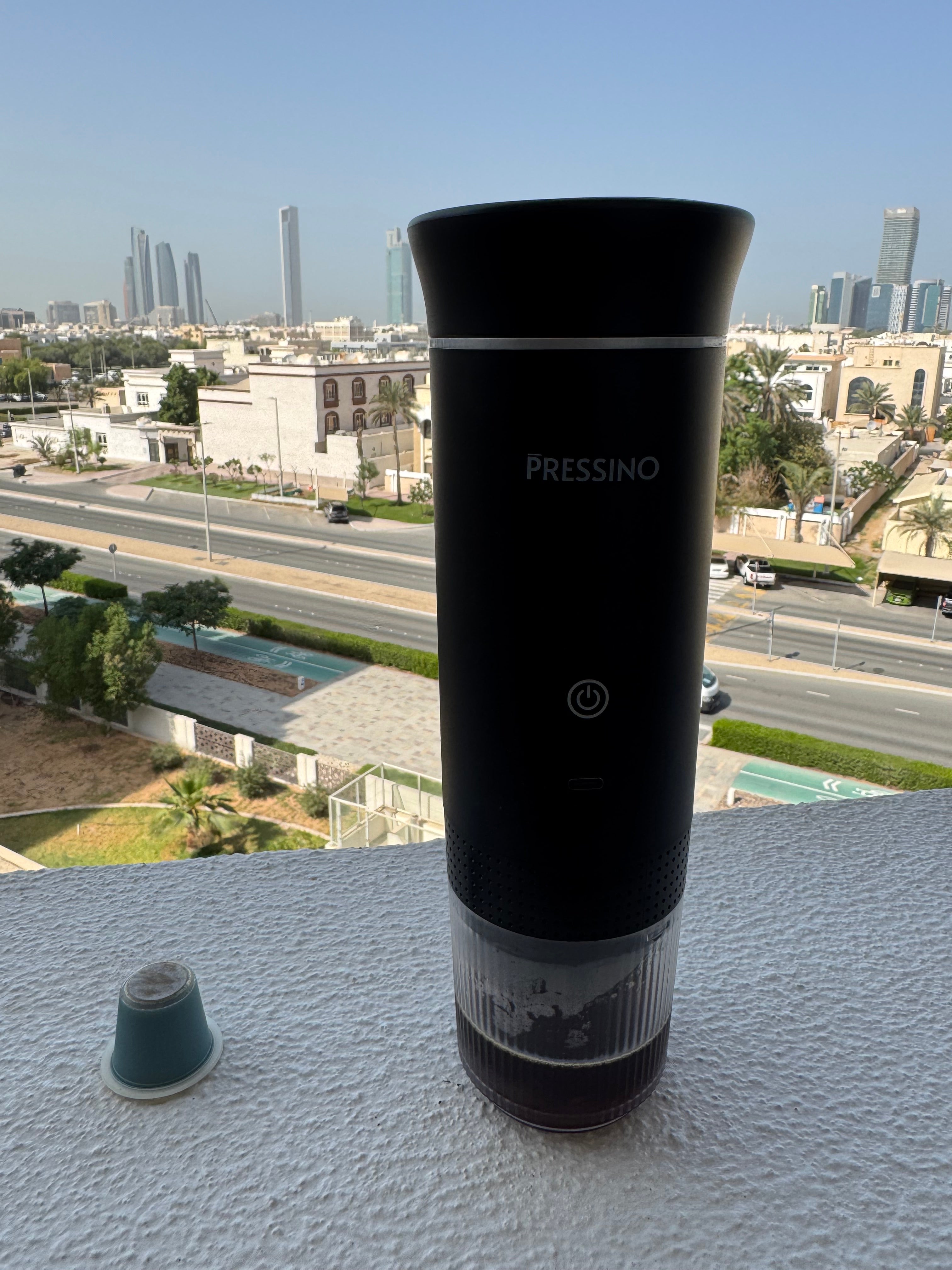 A sleek black portable espresso maker labeled “PRESSINO” stands on a balcony ledge with a city skyline in the background, sunlight highlighting its matte finish, capturing an outdoor coffee moment that blends urban lifestyle with on-the-go espresso craftsmanship.