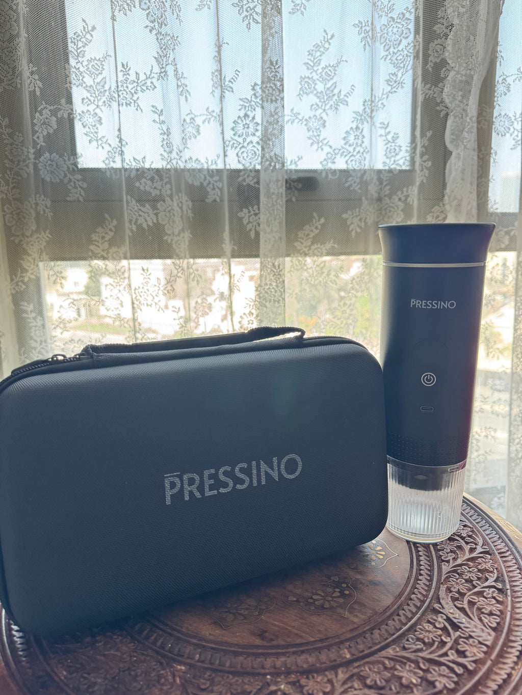 Matte black PRESSINO travel case and skincare-style espresso device displayed on a carved wooden surface by a sunlit window with lace curtains, showcasing elegant minimal design and portability.