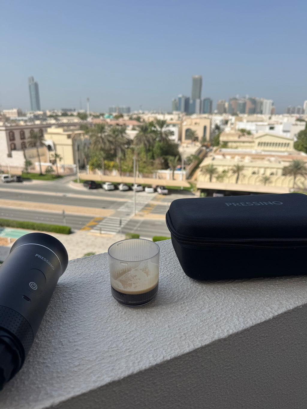 Pressino One portable espresso maker, travel case, and a cup of espresso resting on a balcony ledge overlooking a bright cityscape, capturing the essence of outdoor coffee moments and urban freedom.