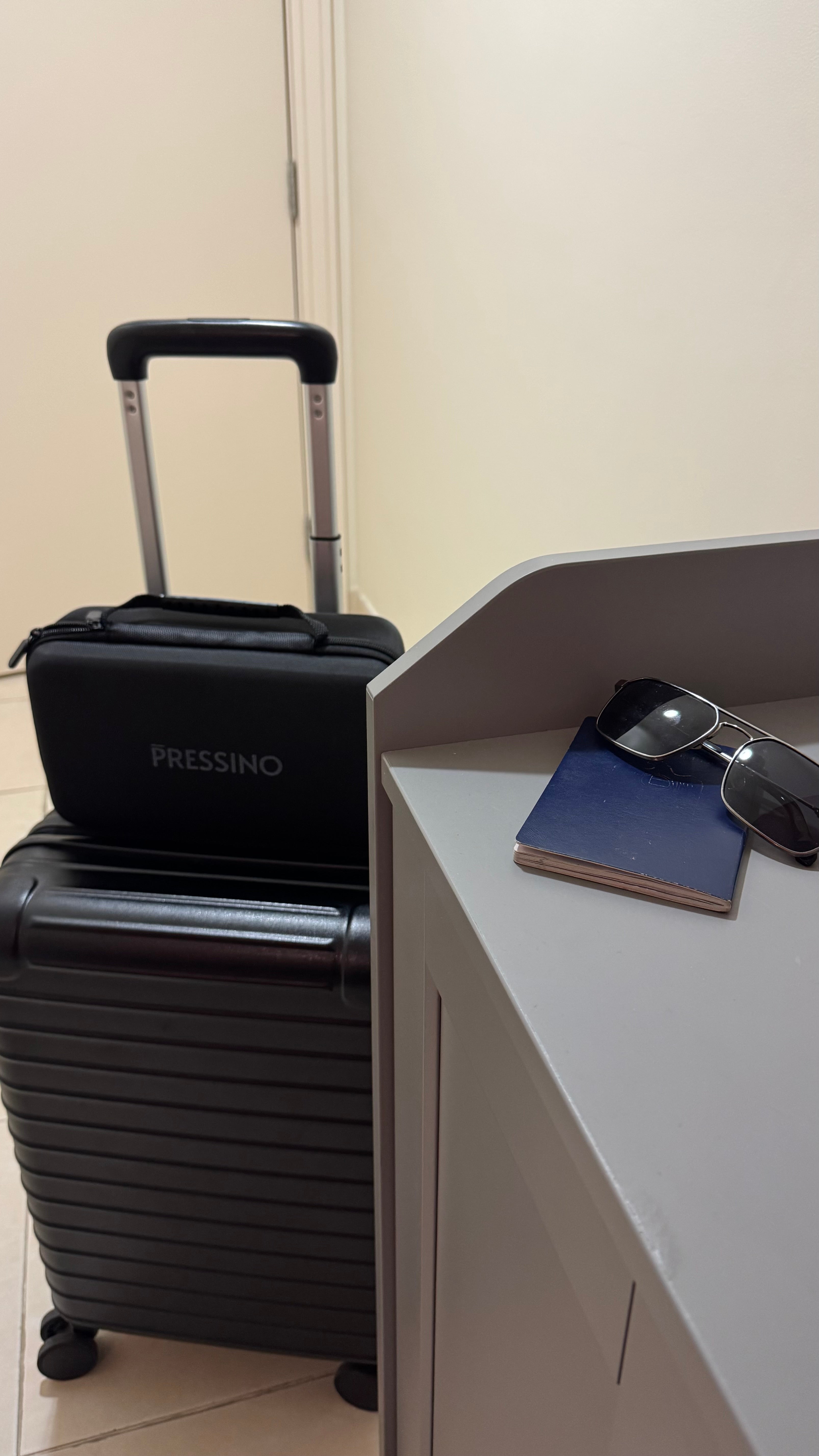 Black Pressino One travel case stacked on a sleek black suitcase beside sunglasses and a blue passport on a white surface, evoking a premium travel aesthetic and on-the-go espresso lifestyle.