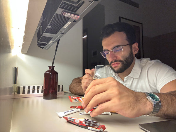 A man wearing glasses and a white polo shirt sits at a lit desk, holding a glass cup while preparing coffee. Several sugar packets and coffee sachets are scattered in front of him, with a dark red bottle and books in the background. The warm lighting creates a focused, calm atmosphere.