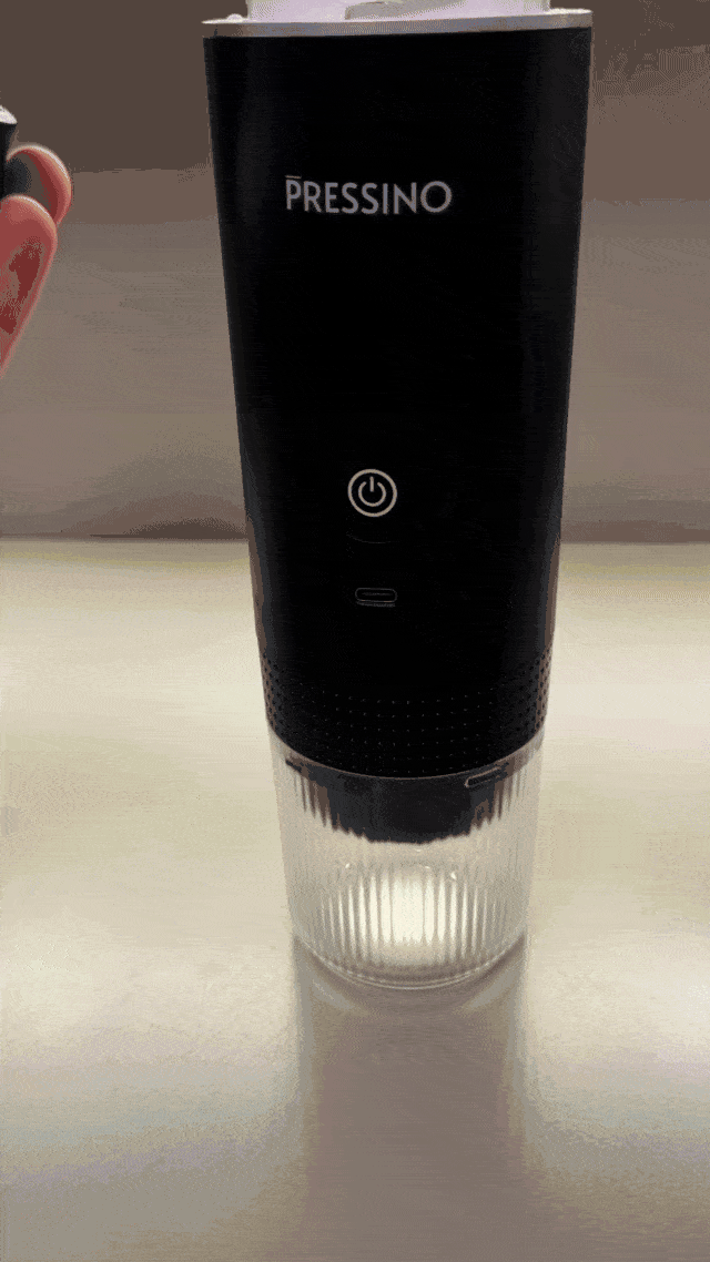 A person installs a coffee capsule into a sleek black portable espresso maker labeled “PRESSINO.” The device sits on a white countertop with a transparent cup attached to its base, demonstrating the first step of preparing espresso using the Pressino machine.
