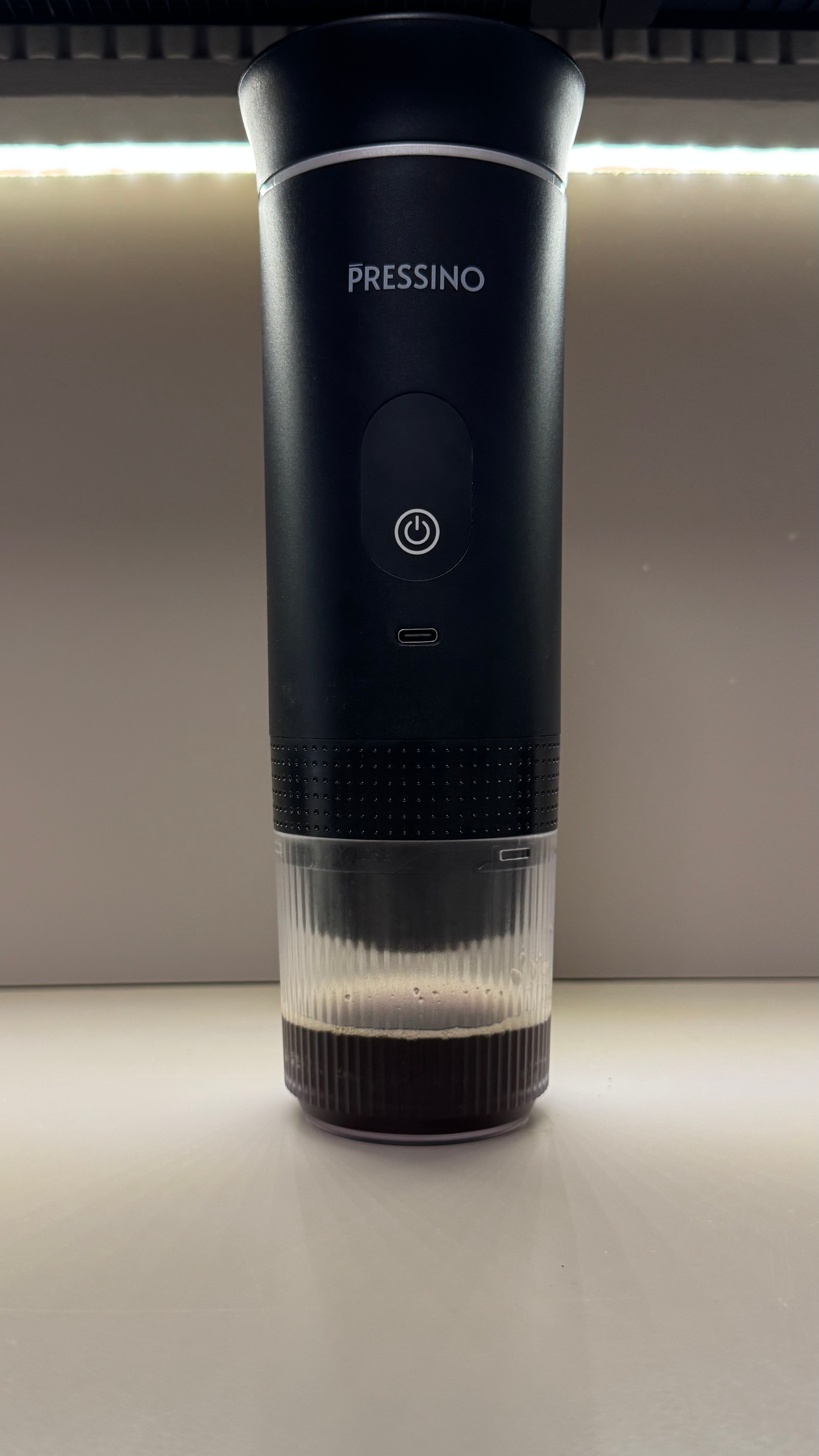 A sleek black portable espresso maker labeled “PRESSINO” sits on a countertop with freshly brewed espresso visible in the transparent cup attached at the base, illuminated under warm lighting.